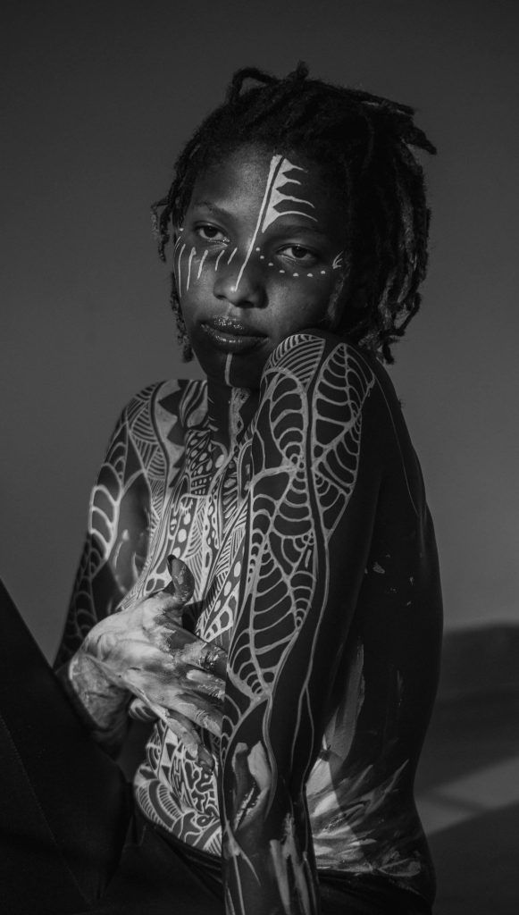 A black and white photo of a person with intricate body and face paint, adorned with tribal tattoos, wearing their hair in short twists and sitting with one arm across their chest. The image captures the cultural significance and stories behind the ink on their skin.