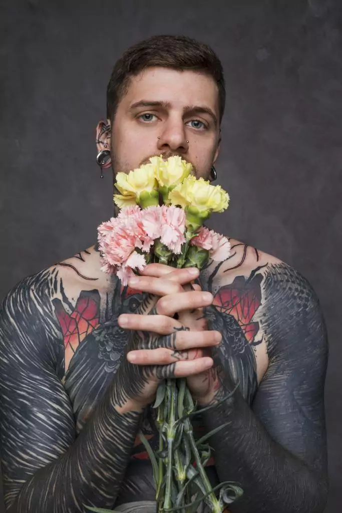A person with extensive San Diego tattoos holds a bouquet of yellow and pink flowers against a dark background.
