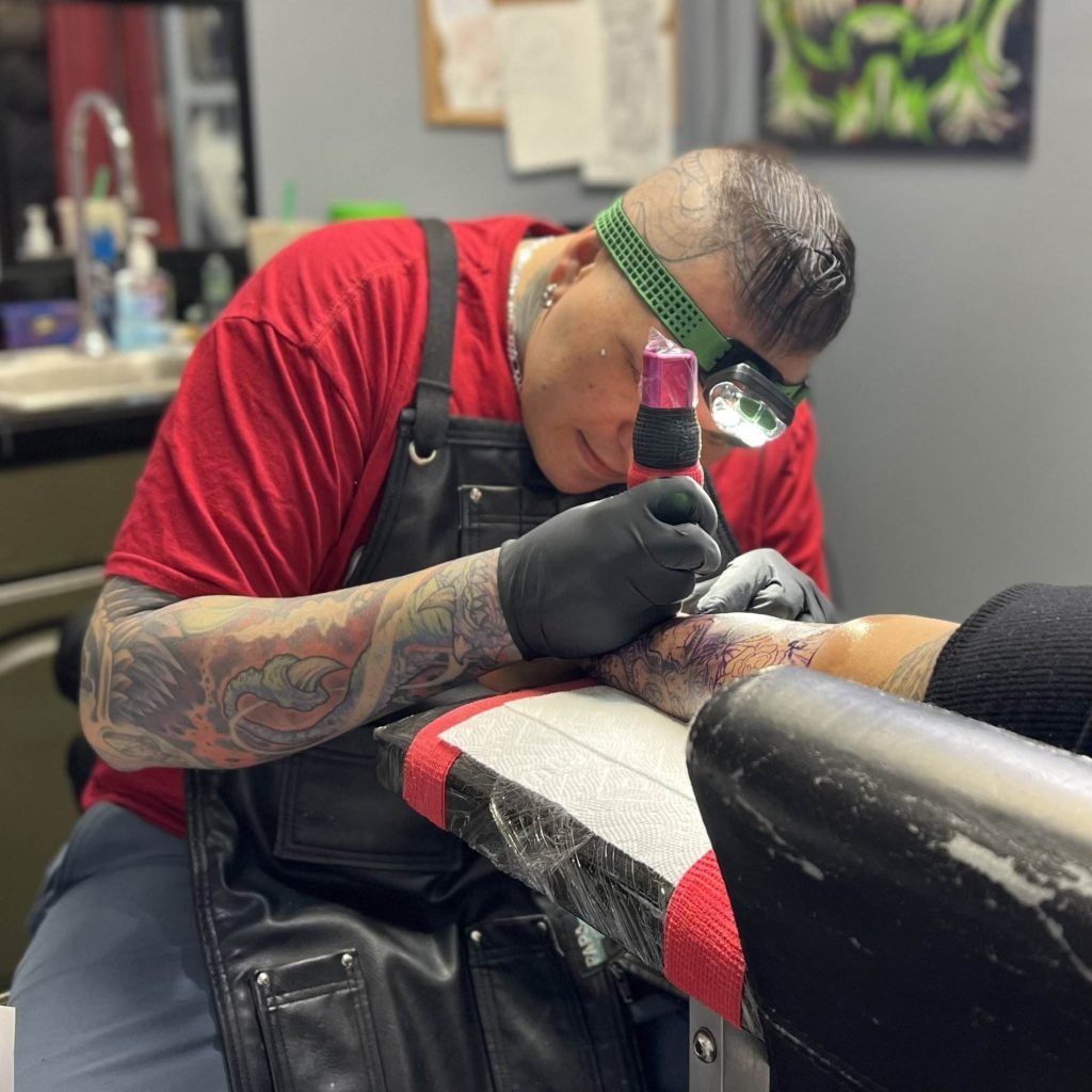 Tattoo artist in a red shirt and black apron, wearing a headlamp, meticulously tattoos a client's arm with a Zorac design on a padded table in the tattoo shop.