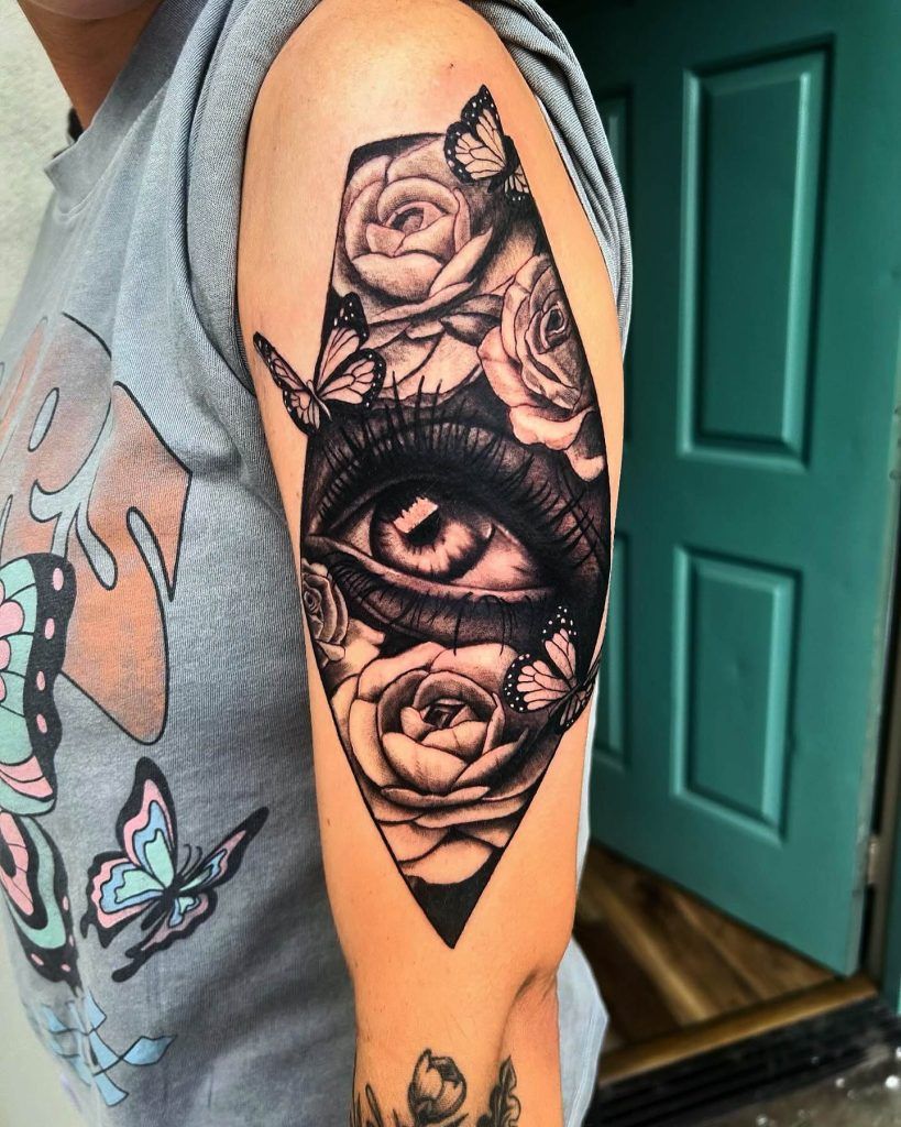 A detailed tattoo featuring a realistic eye surrounded by roses and butterflies on a person’s upper arm.