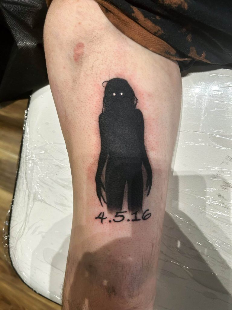 A black silhouette of a person with white eyes is tattooed on an arm above the date "4.5.16." The tattooed arm is resting on a white surface.