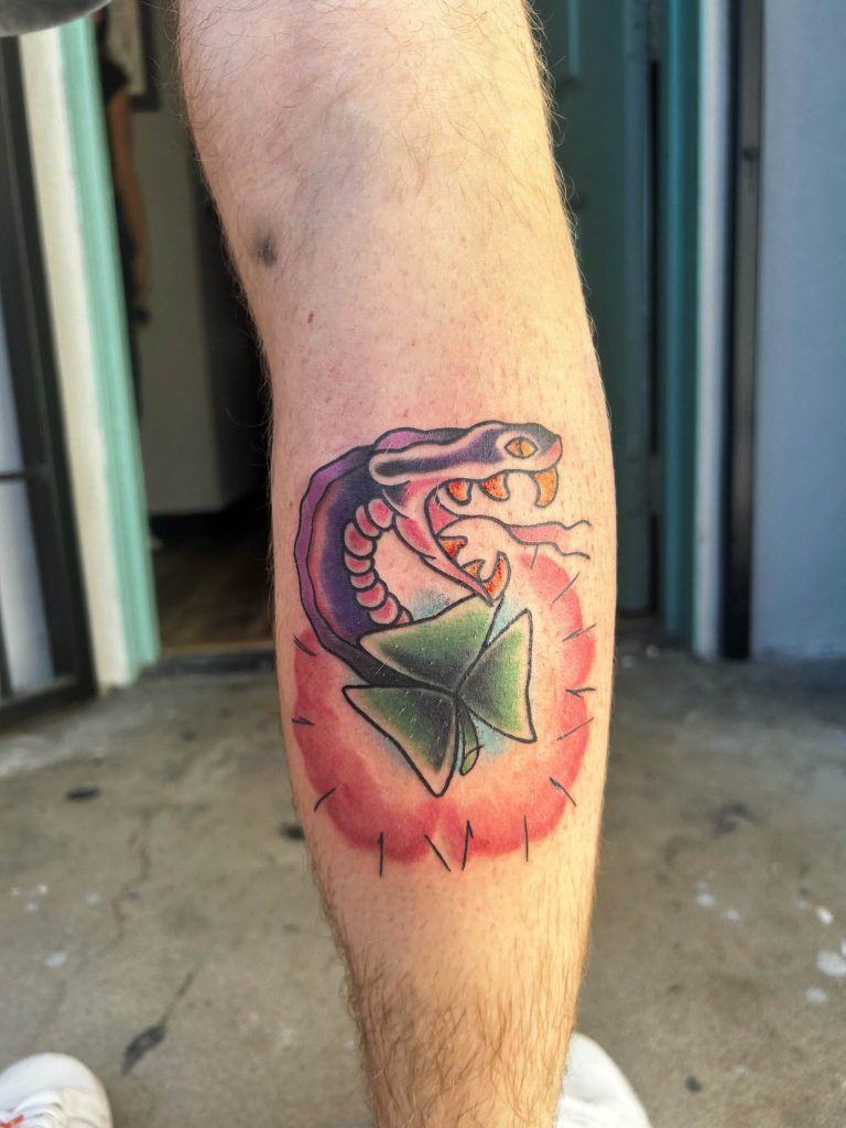 A leg tattoo featuring a purple and black snake coiled around a green bow within a red circular background.