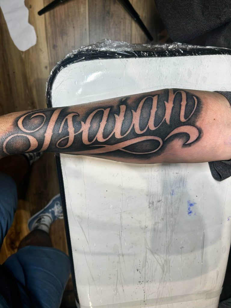 Forearm tattoo showing the name "Isaiah" in large, elaborate script. The background includes a partially visible person and art studio equipment on a wooden floor.