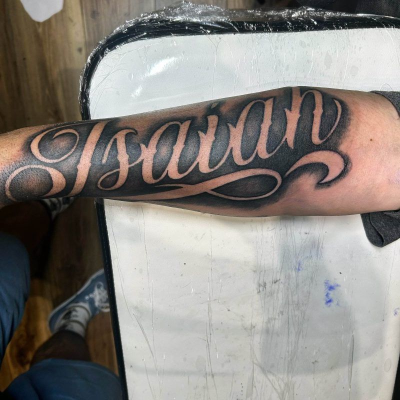 Forearm tattoo showing the name "Isaiah" in large, elaborate script. The background includes a partially visible person and art studio equipment on a wooden floor.