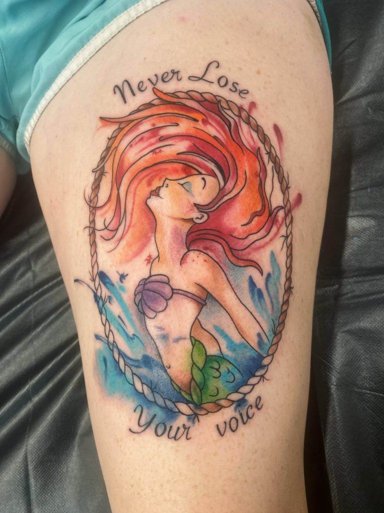 Colorful tattoo of a mermaid with flowing red hair and a purple top encircled by a rope with the text "Never Lose Your Voice" on a person's leg.