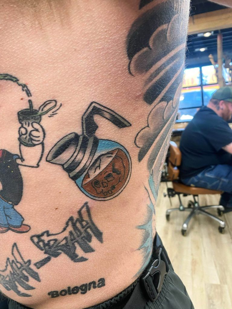 Tattoo of a coffee pot with the word "Death" on the pot, surrounded by other tattoos, on a person's body. A person is visible in the background sitting inside a room.