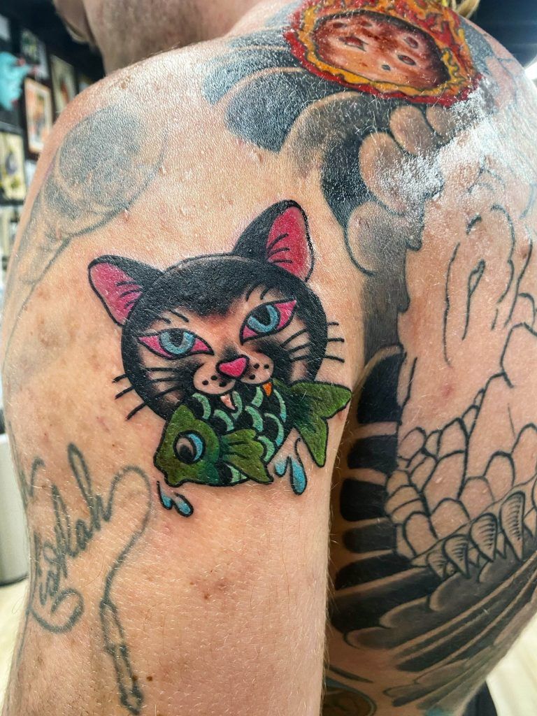 A person has a tattoo on their shoulder depicting a black cat with pink ears holding a green fish in its mouth, surrounded by other tattoos.