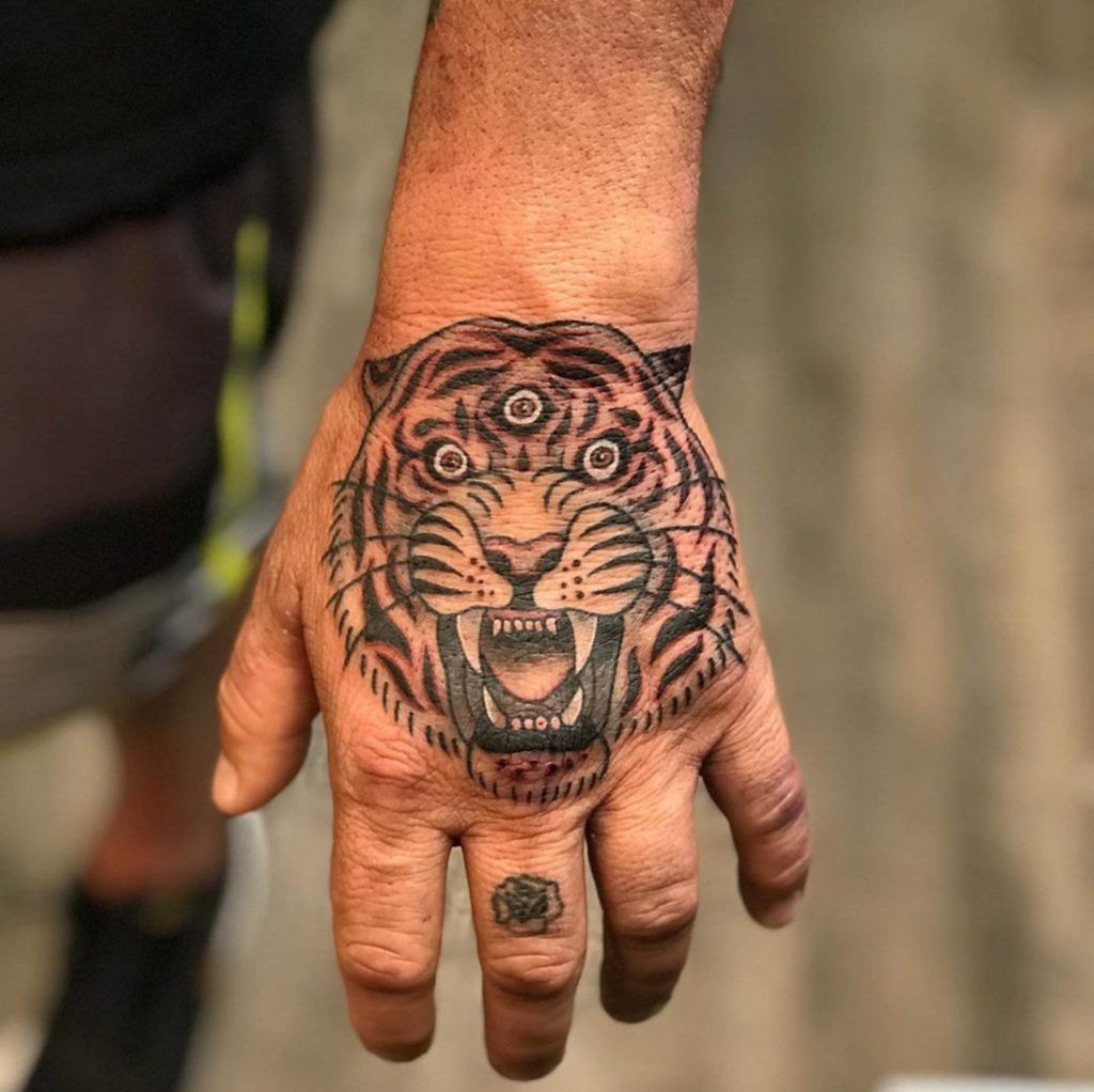 A tattooed hand featuring a detailed tiger's face with three eyes on the back of the hand, embodying the mysteries of tattoo symbolism, and a smaller flower tattoo on the ring finger.