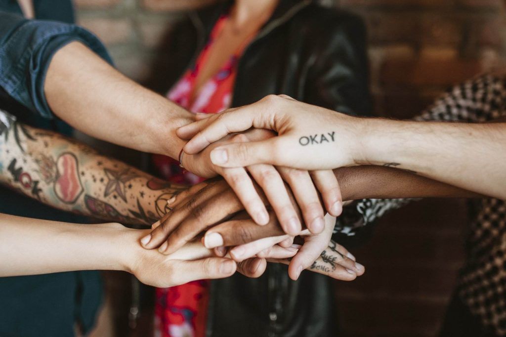 A group of people place their hands together in a stack. Some hands have tattoos, with one displaying the word "OKAY." They are standing against a blurred brick wall background, showcasing powerful forms of self-expression.