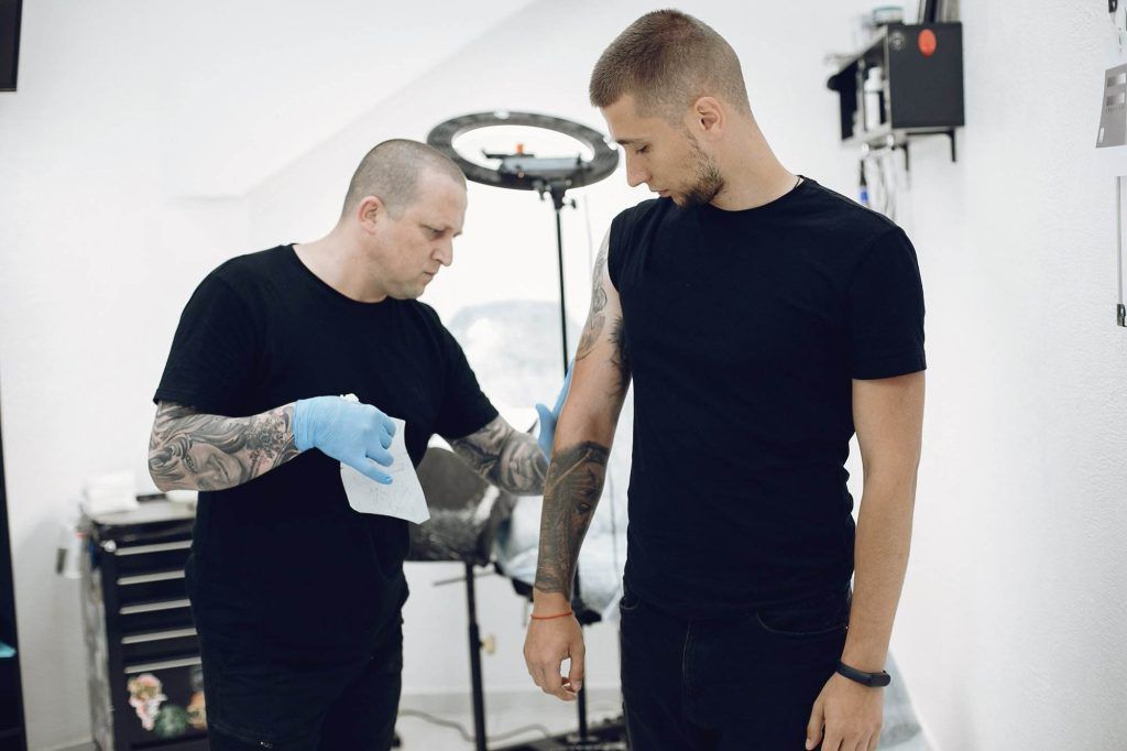 A tattoo artist wearing gloves prepares to work on a client's arm tattoo in a studio. The client stands next to him, both in black clothing, setting the scene for an intricate step-by-step guide to the perfect ink.