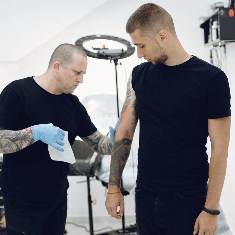 A tattoo artist wearing gloves prepares to work on a client's arm tattoo in a studio. The client stands next to him, both in black clothing, setting the scene for an intricate step-by-step guide to the perfect ink.