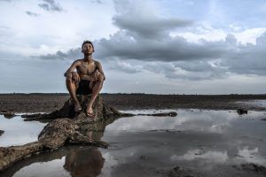 A shirtless person, adorned with popular tattoos, sits on a tree stump in a barren, muddy landscape under a cloudy sky.