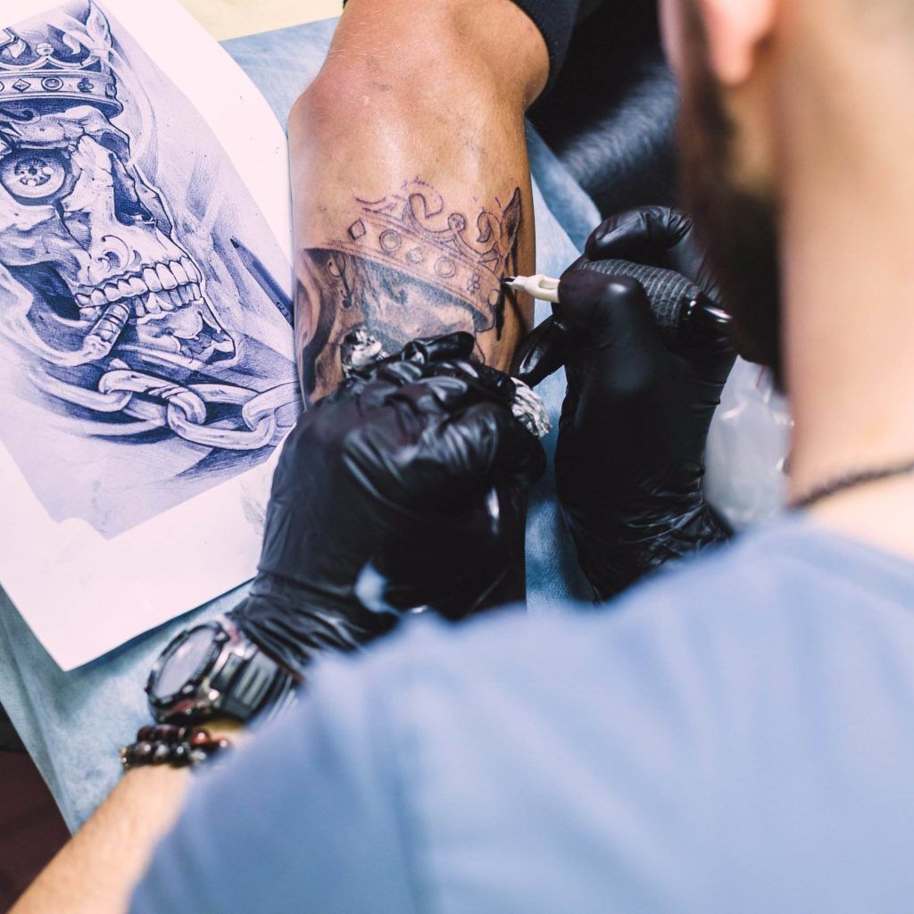 A tattoo artist wearing black gloves is working on a vibrant skull with a crown on a person's arm, carefully blending in the color pigments. A reference image of the skull tattoo is placed nearby.