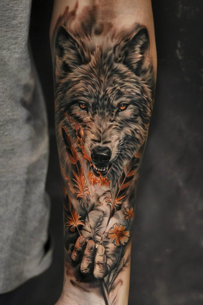 A detailed tattoo of a wolf's head with piercing eyes, intertwined with realistic orange feathers and flowers, covers the entire forearm of a person, embodying popular tattoo designs rich in symbolism.