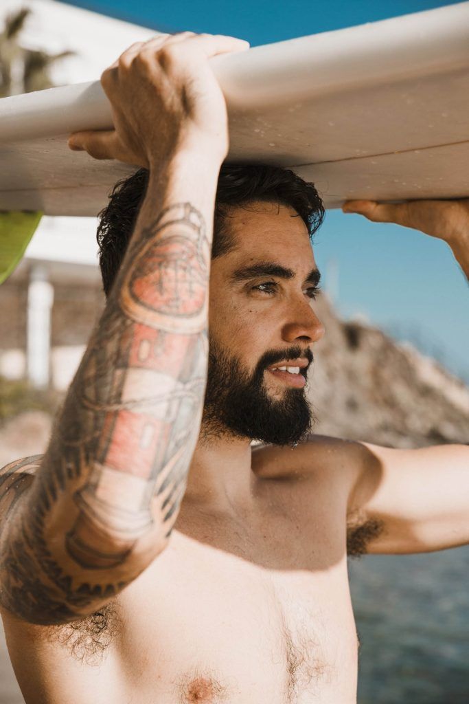 A shirtless man with a beard and arm tattoos holds a surfboard above his head on a sunny day at the beach, showcasing how to age with grace while preserving the beauty of an active lifestyle.