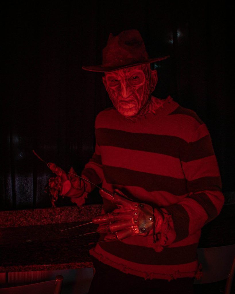 Person in a detailed costume with a striped sweater, wearing a fedora, a glove with blade-like extensions, and heavy makeup under red lighting, resembles one of horror's iconic characters. Tattoos peek out from beneath the sleeves, adding an extra layer of intrigue.