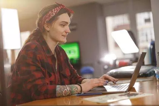 In a bright room, a person with visible tattoos and a red bandana embodies the 2025 workplace attitudes as they navigate remote work culture, using a laptop at a wooden table.