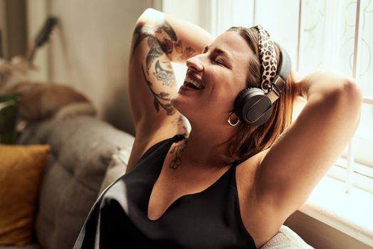A woman with intricate skin art relaxes on a couch, wearing headphones and smiling with eyes closed, as though savoring the whispers of distant voices.