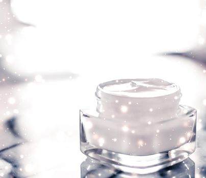 A jar of white cream sits elegantly on a reflective surface with a blurred, luminous background, ready to enhance your seasonal tattoo routine. Specks of light dance around the jar, promising nourishment and care for vibrant skin art.