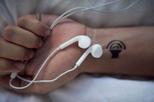 A hand holding white earphones, with a tattoo resembling a sound wave on the wrist—a tribute to music and the voices it carries.