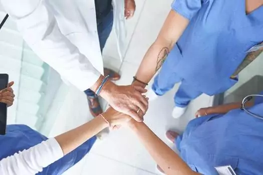 Healthcare professionals in scrubs, some with visible tattoos, join hands in a circle, symbolizing teamwork.