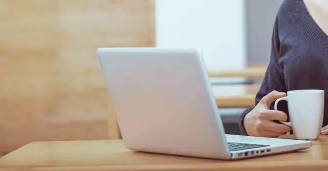 A person engaged in remote work culture sits at a table with a laptop, holding a white mug.