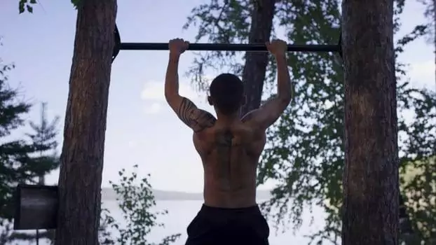 A tattooed individual embraces an active lifestyle, performing pull-ups on a bar between two trees, set against the scenic backdrop of a tranquil lake and lush forest.