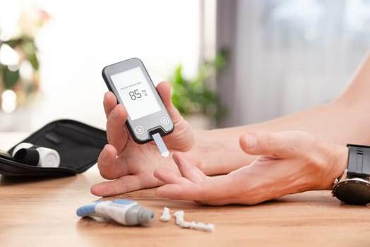 A person, tattooed with diabetes awareness symbols, checks blood sugar with a glucometer at a table. The reading shows 85, following 2025 medical guidelines. Nearby, a case and accessories lie neatly arranged.