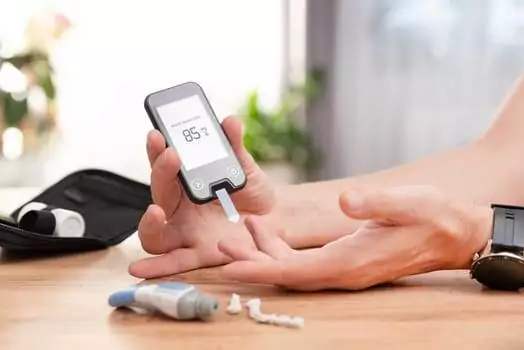 A person, tattooed with diabetes awareness symbols, checks blood sugar with a glucometer at a table. The reading shows 85, following 2025 medical guidelines. Nearby, a case and accessories lie neatly arranged.
