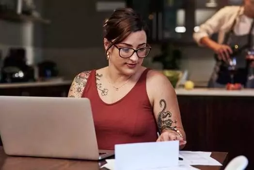 A person with glasses and visible tattoos sits at a table by a laptop, embodying the modern remote work culture, while looking at a piece of paper. In the background, someone else is busy in the kitchen.
