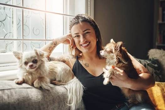A person smiles while sitting on a couch with two small dogs, one on their lap and the other beside them. Sunlight streams through a window in the background, casting a warm glow reminiscent of cherished pet memorial tattoos captured forever in heart and ink.