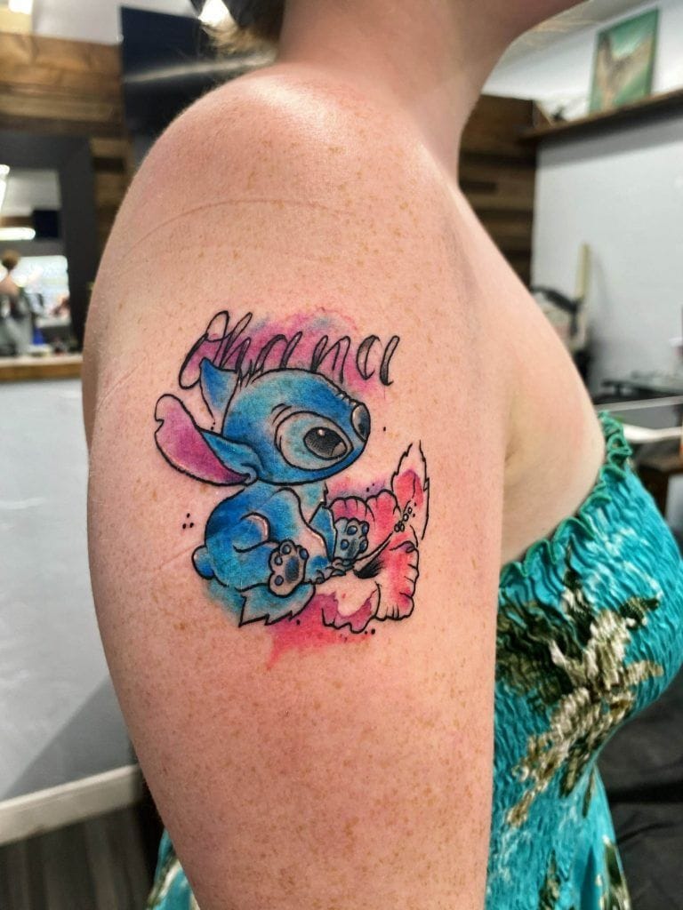 Tattoo of a blue cartoon character holding a pink flower on someone's upper arm, with the word "Ohana" above it.