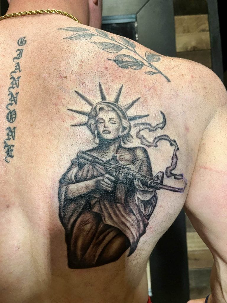 Back tattoo depicting the Statue of Liberty holding a rifle, with leaves above. The word "GANNON" is tattooed vertically on the left side.