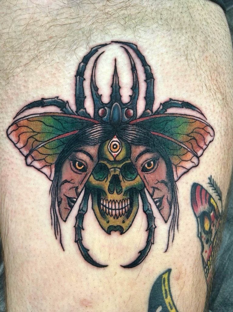 Tattoo of a beetle with colorful wings, featuring a central skull and two faces on each side.