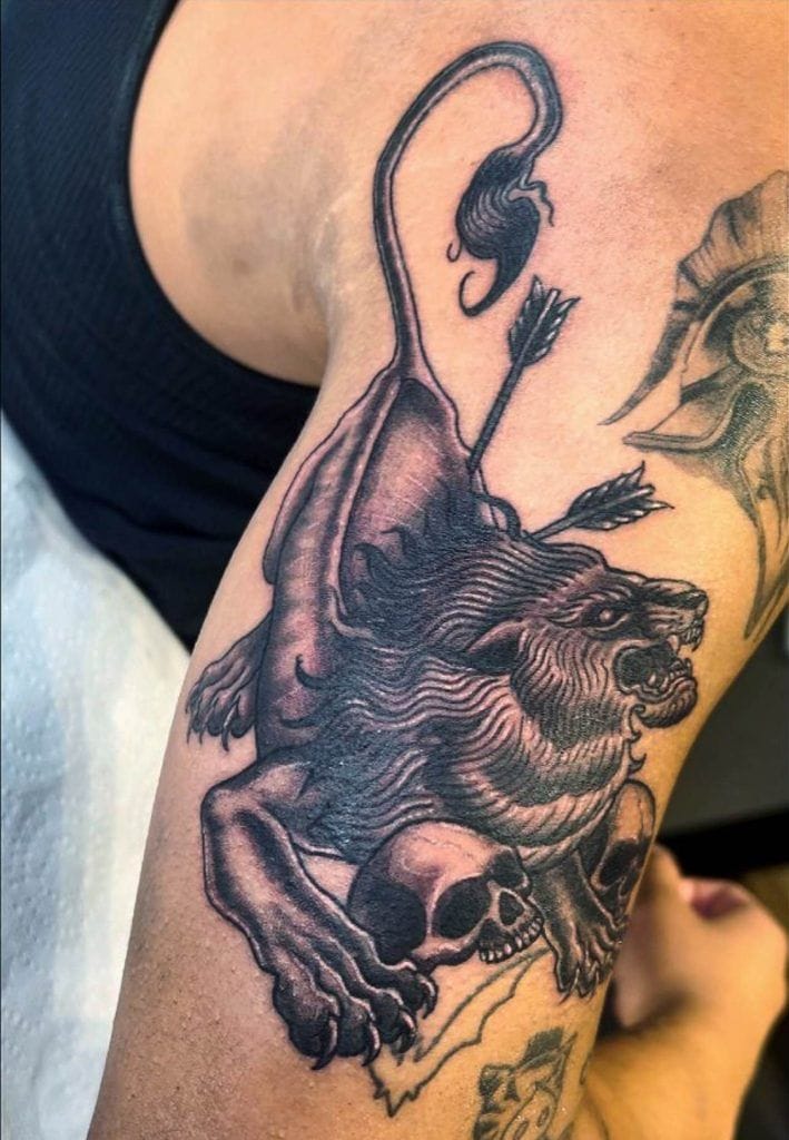 Tattoo of a roaring lion with arrows and a skull on an upper arm.