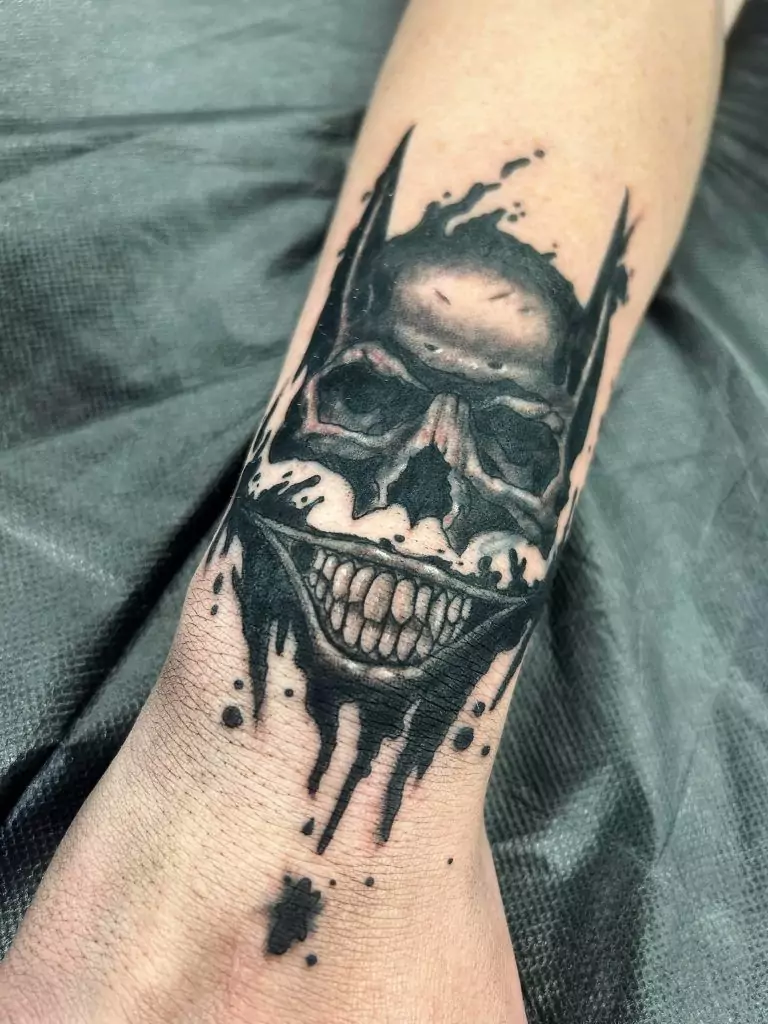 Forearm tattoo of a grinning skull with bat-like ears and ink splatters, on a textured dark background.