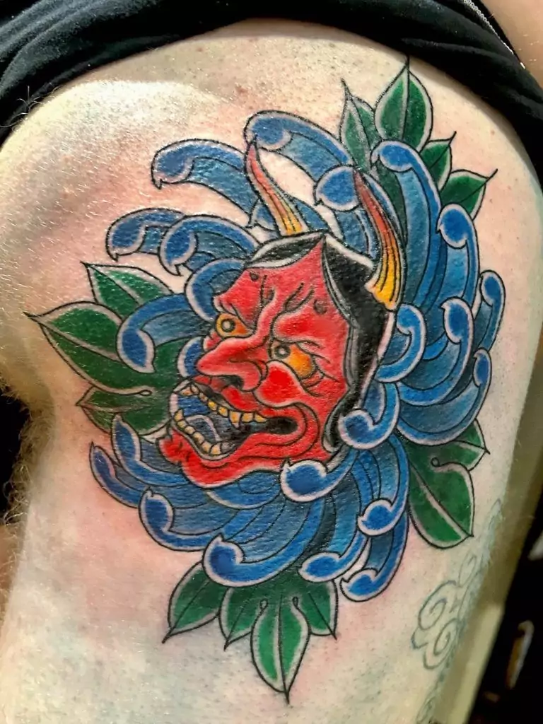 Tattoo of a red demon mask with horns, surrounded by blue waves and green leaves on a person's upper arm.