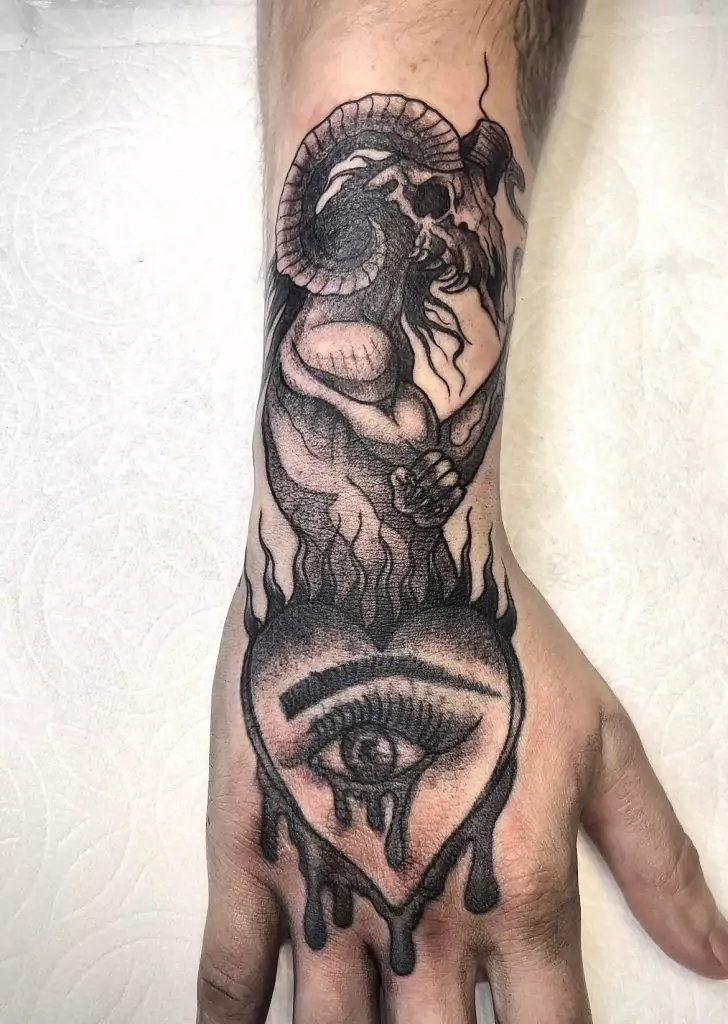 A tattoo on a forearm depicting a horned figure above a large eye with tears and flame-like details on the hand.