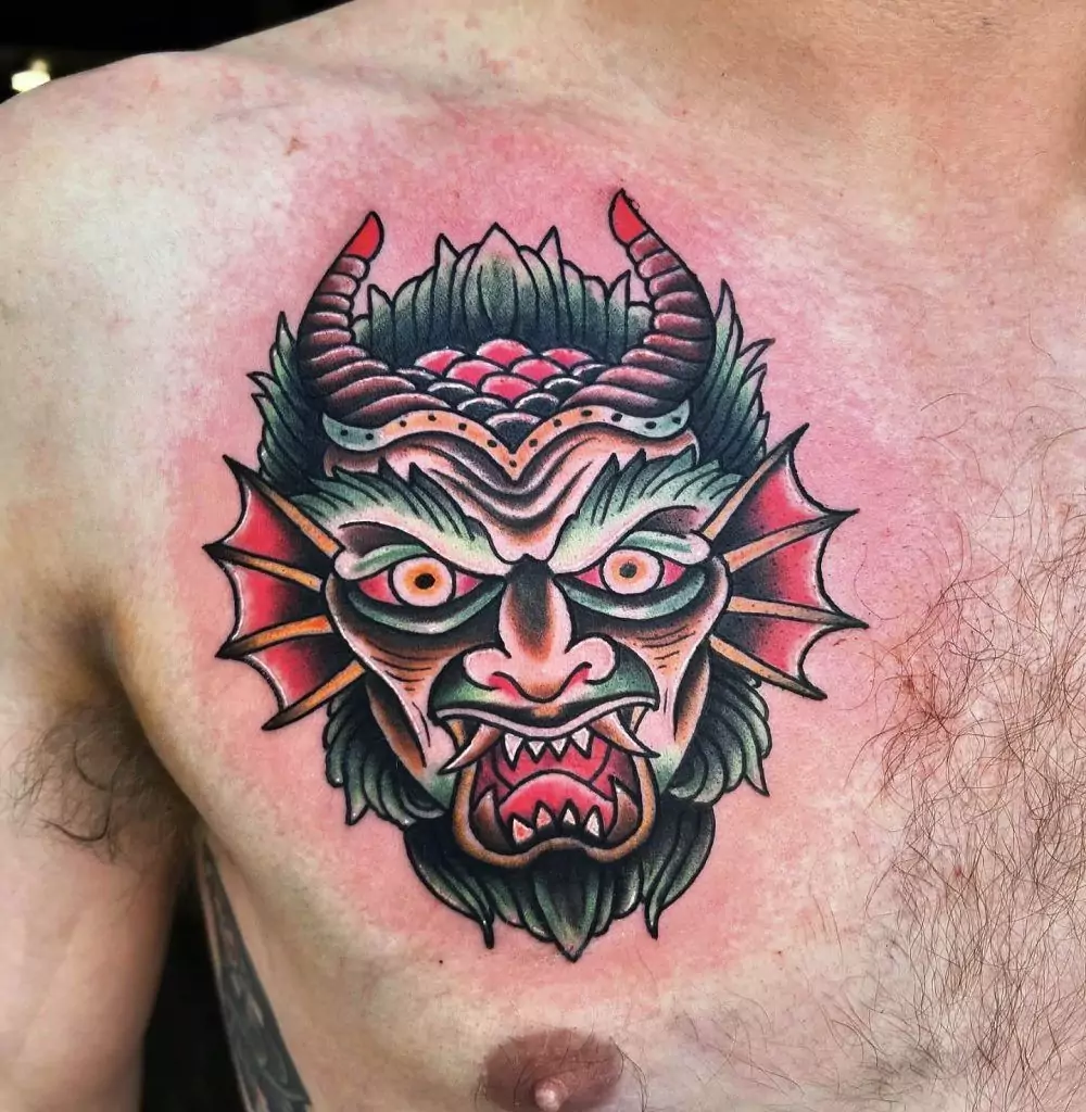 Colorful demon face tattoo on a person's chest with horns, sharp teeth, and an intense expression.