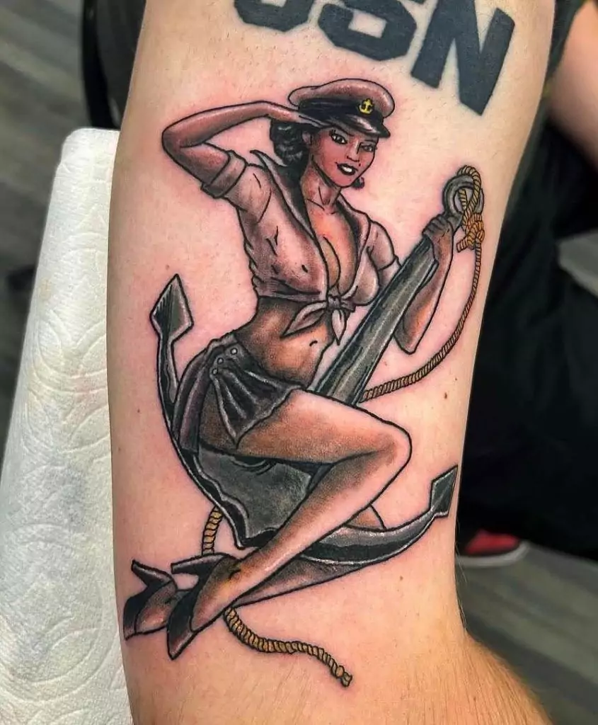 Tattoo of a pin-up sailor woman sitting on an anchor, with a rope, wearing a sailor hat and an open collared shirt.