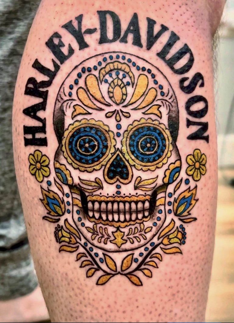 Colorful sugar skull tattoo with "Harley-Davidson" text on skin, featuring floral and geometric designs in blue, yellow, and black.