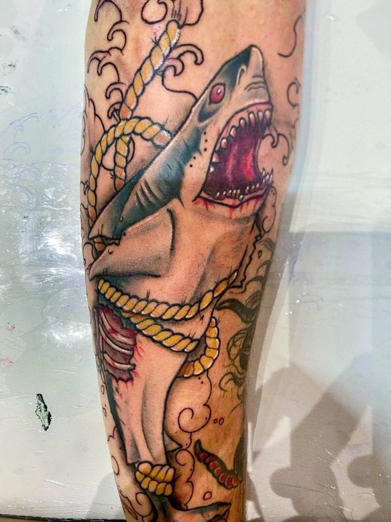 A colorful tattoo of a shark wrapped in yellow rope, with an open mouth and detailed shading, on a person's arm.