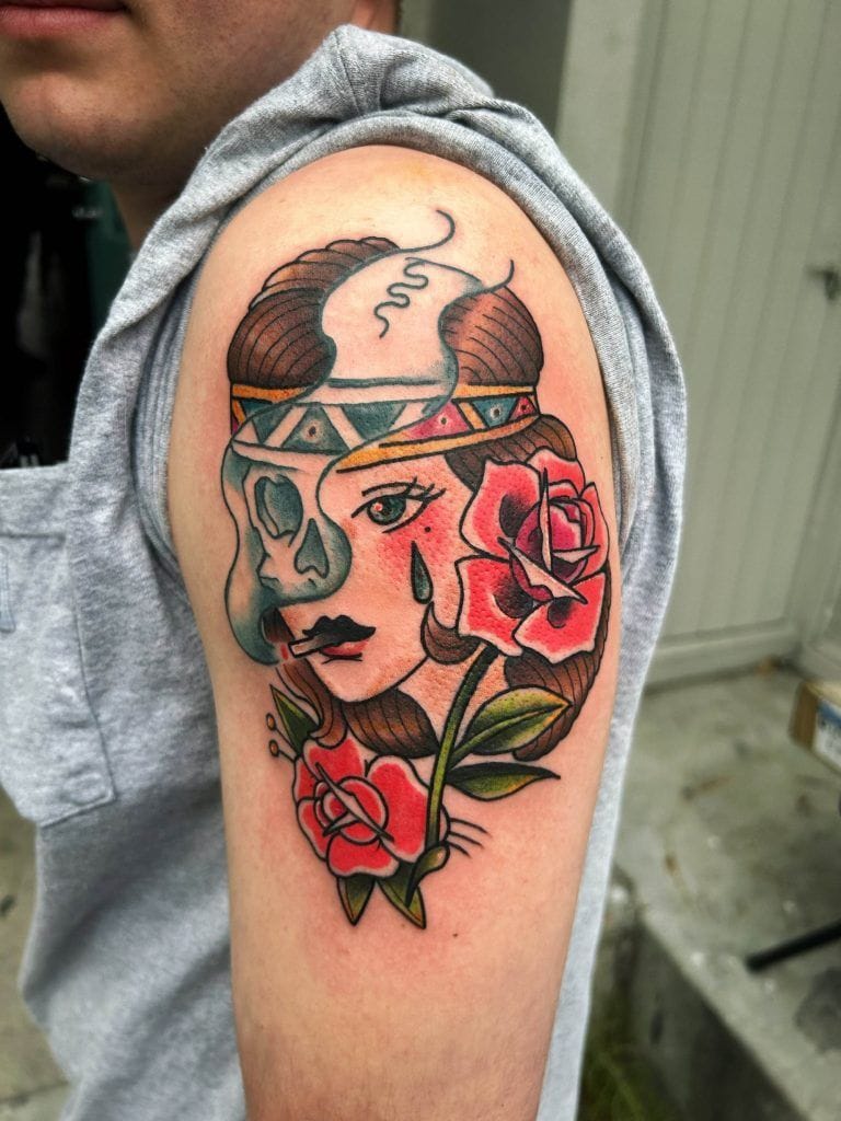 Tattoo of a woman's face with tribal face paint and a skull on her head, surrounded by red roses on a person's upper arm.