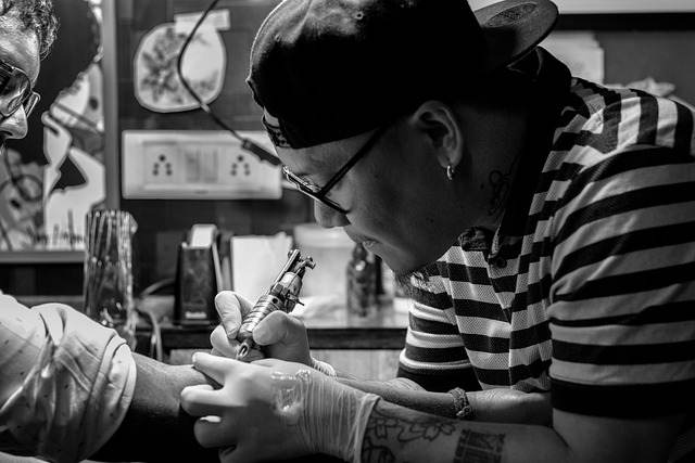 In a studio, a tattoo artist in a striped shirt and cap focuses intently while ink flows onto the client's arm, ensuring quality that defies the notion of cheap tattoos.