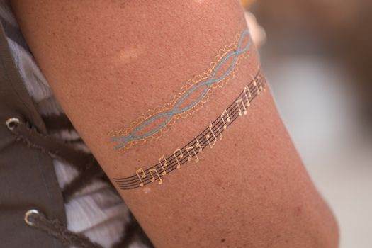 An arm adorned with two temporary tattoos: one showcasing blue and gold intertwined patterns and the other capturing the essence of music with a musical staff, notes, and echoes of sound wave tattoos.