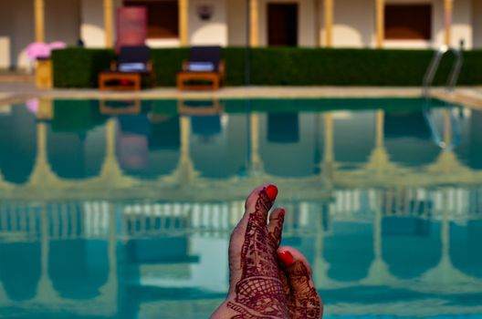 Feet adorned with henna rest near a shimmering pool, where two lounge chairs invite relaxation and safety guidelines whisper in the background.
