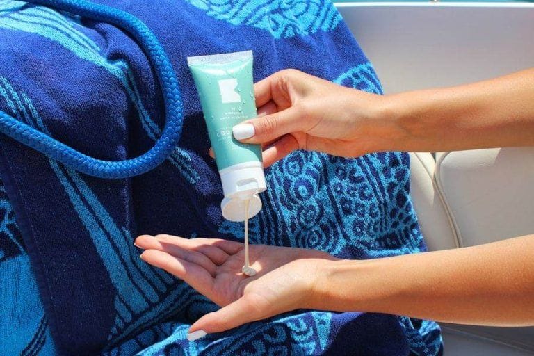 Person squeezing sunscreen from a tube onto their hand, mindful of tattoo aftercare. A blue patterned towel lies in the background, adding a splash of color to this sun-soaked scene.