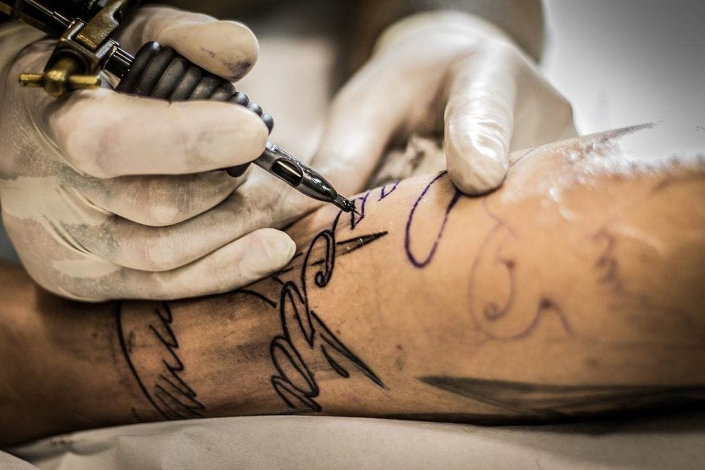 A tattoo artist wearing gloves deftly uses a tattoo machine to ink a design on a person's arm, showcasing different styles that ensure the artwork remains captivating even after 10+ years.