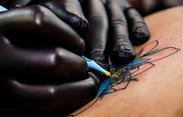 Close-up of a gloved hand using a tattoo machine to ink a blue and yellow flower design on skin, skillfully transforming surgery scars into artful memories with tattoos designed for seamless scar coverage.