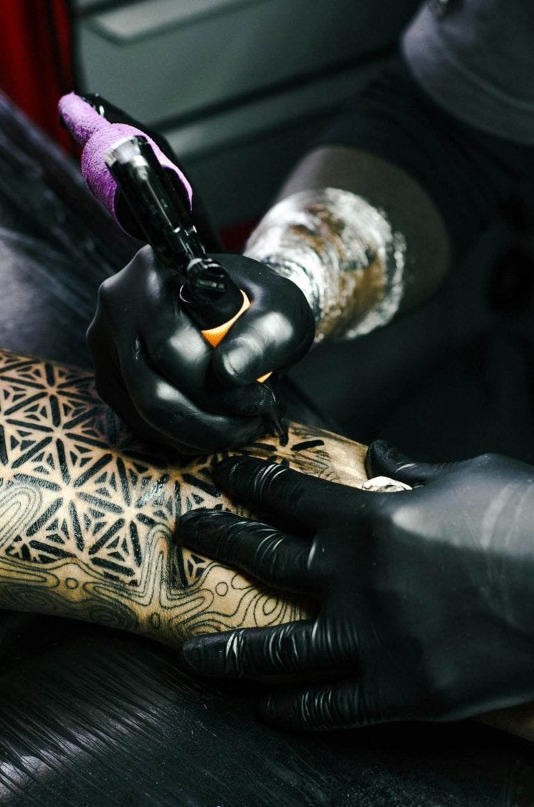 Person with black gloves tattooing a geometric pattern on a persons arm.